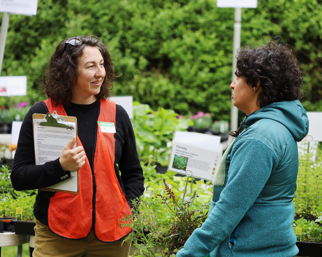 A volunteer in an orange vest holds a clipboard while talking with a customer at a plant sale surrounded by seedlings and educational signs.